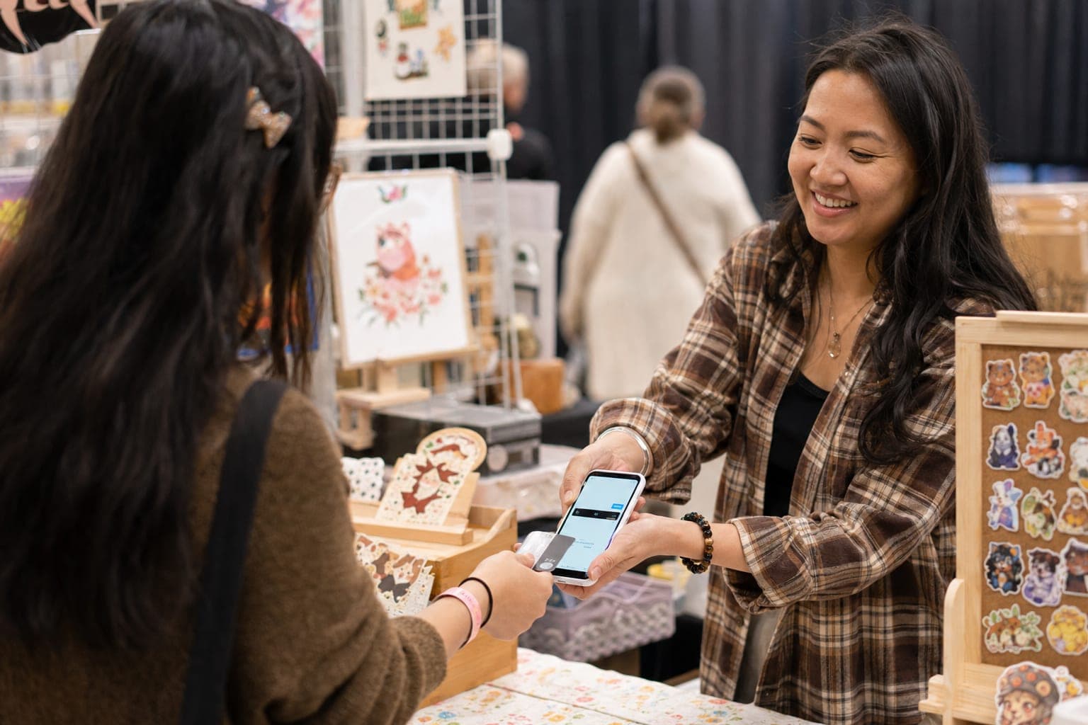A craft fair vendor holding a phone toward a customer's contactless credit card across a handmade-goods booth table