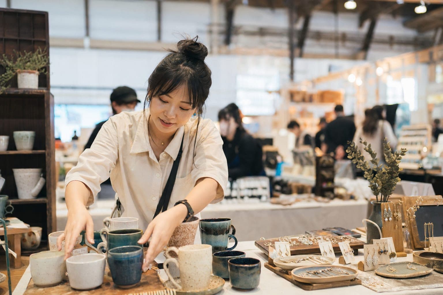 A handmade-goods vendor at a busy indoor craft fair booth arranging ceramic mugs and jewelry on a wooden table, warm daylight, other booths blurred in the background