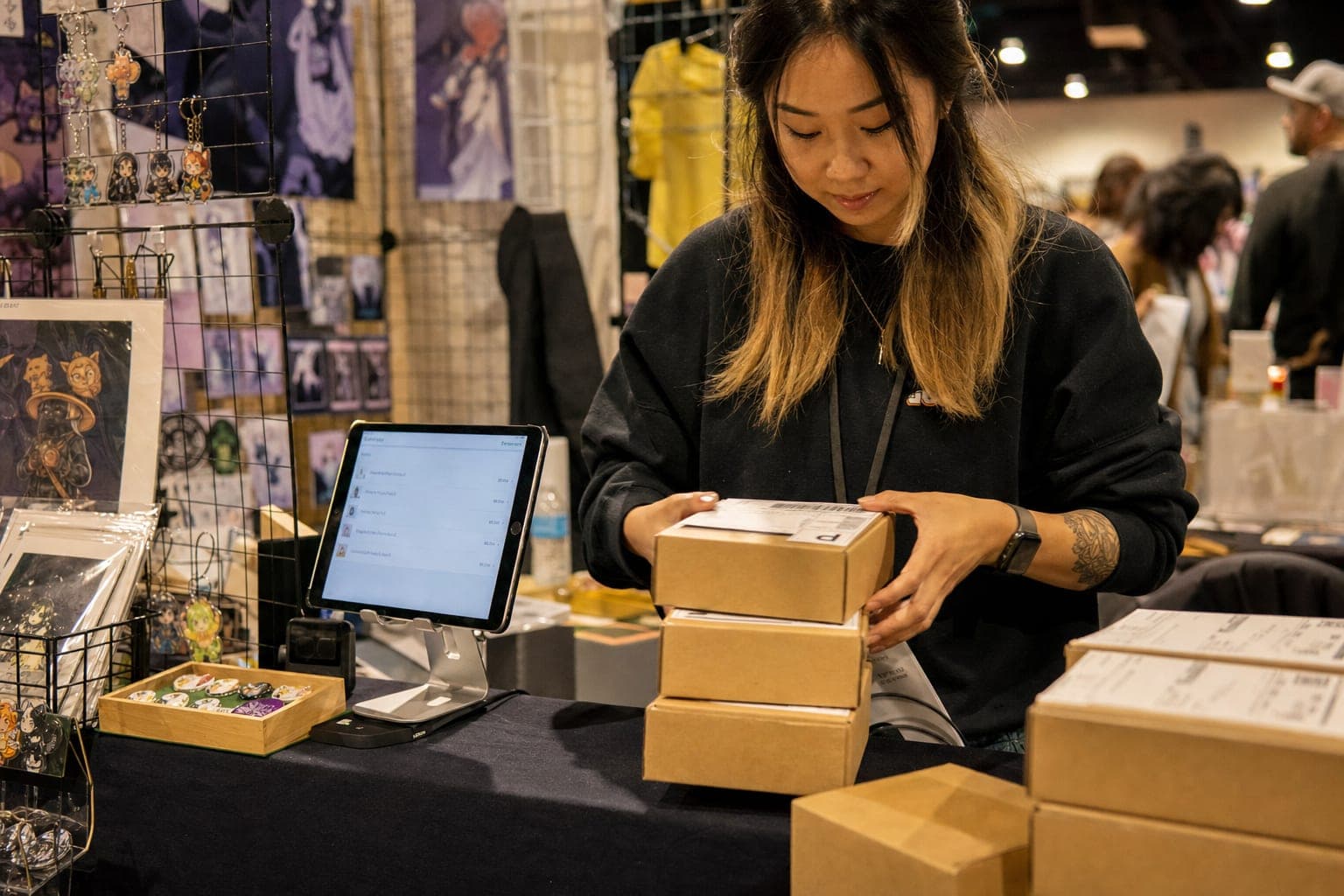 Convention vendor at a craft show booth stacking small brown shipping boxes with printed carrier labels next to a tablet showing live shipping rates