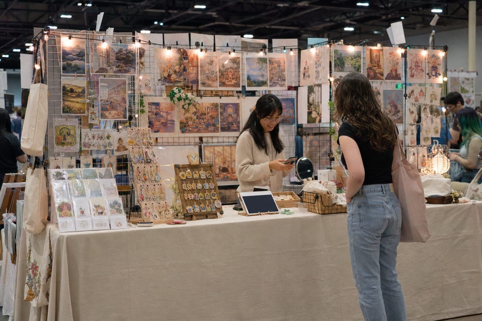 A fully set-up 10x10 craft show booth with a floor-length tablecloth, vertical grid-wall display, string lighting, clear price signs, and an open front aisle