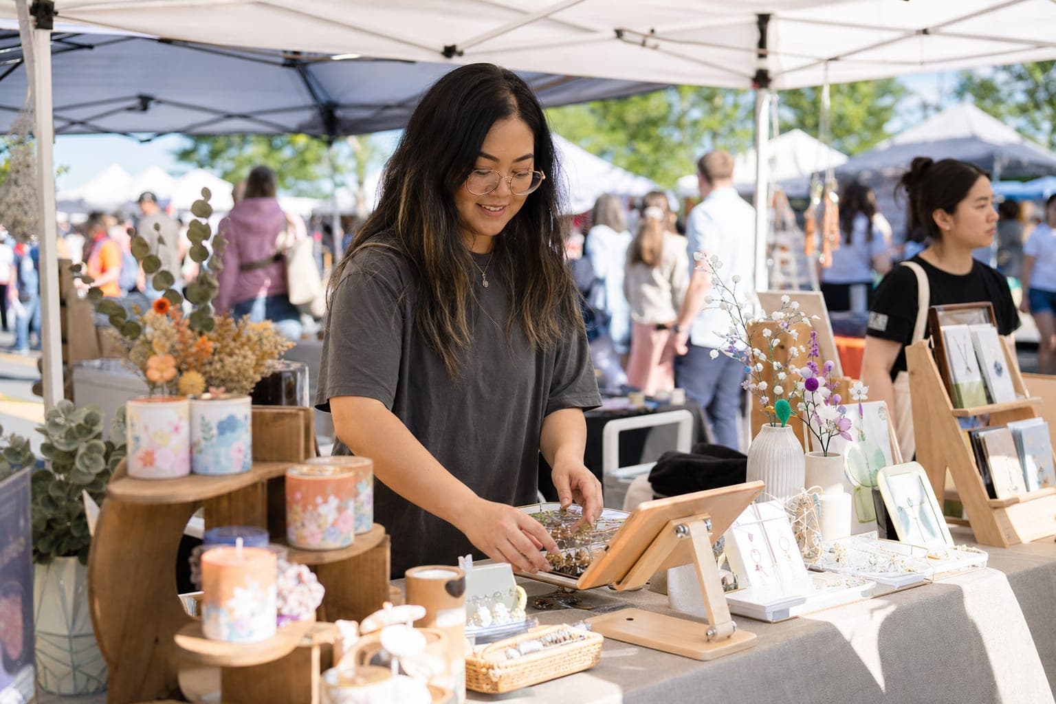 A first-time handmade vendor arranging products on a draped table under a white pop-up tent at an outdoor craft fair, with a tablet propped up for taking orders