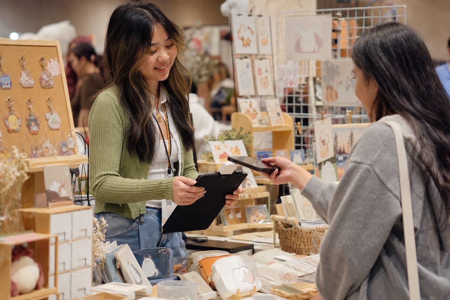 A craft show vendor tapping a customer's phone on a small card reader clipped to a tablet at a booth table stacked with handmade goods